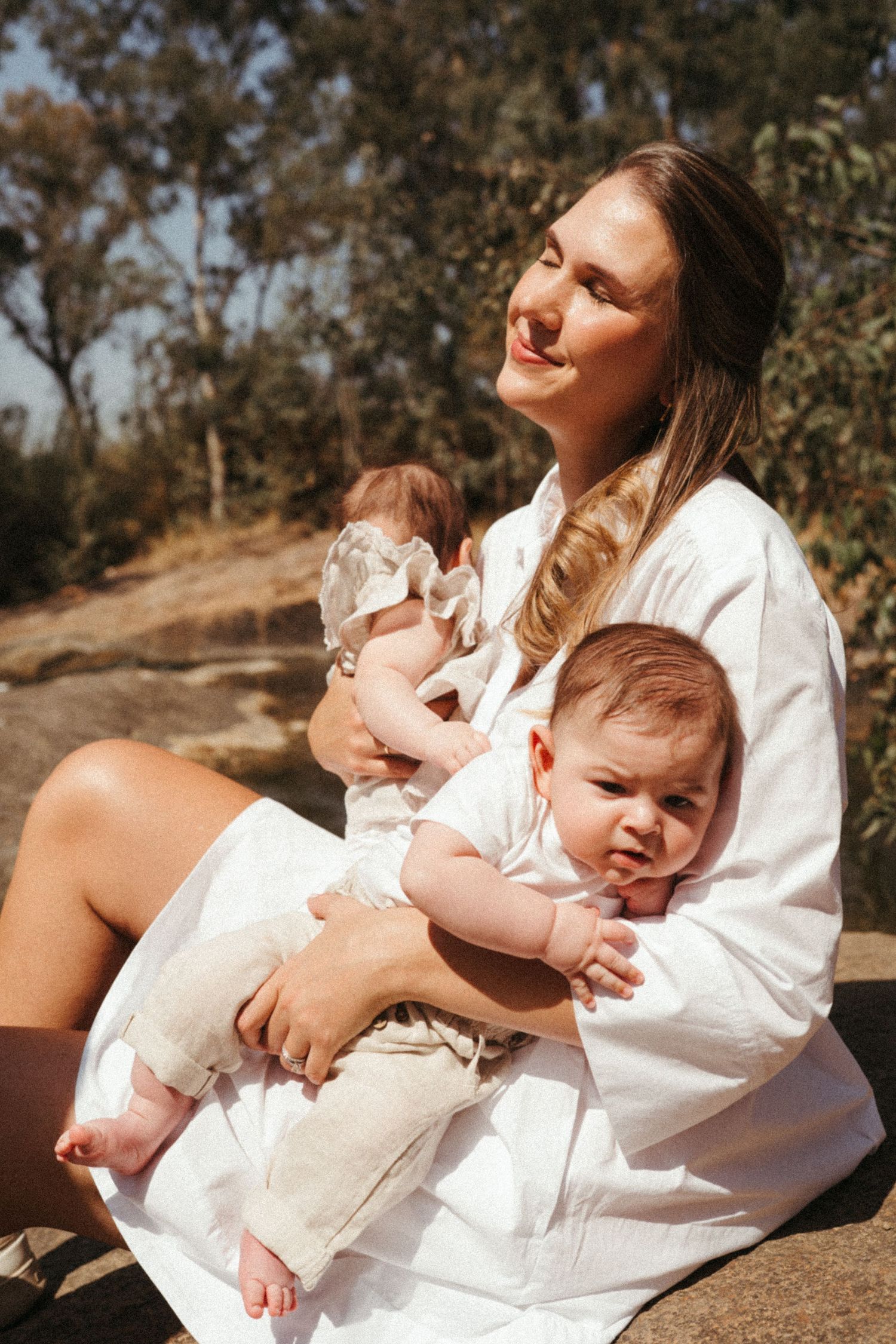 A parent in white holds two infants while sitting outdoors on a rocky surface on a sunny day.