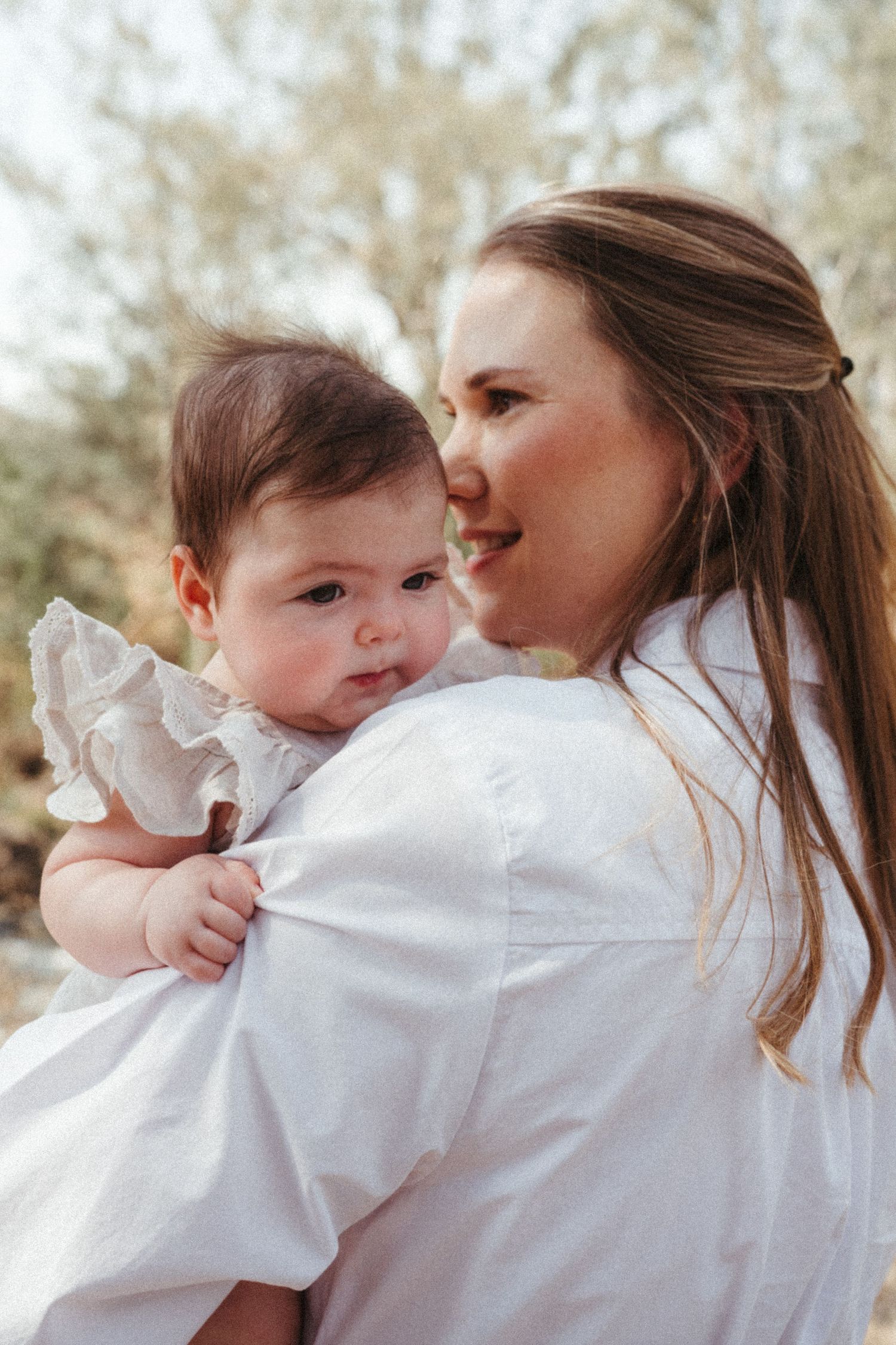 A tender moment between parent and baby in white clothing against a natural outdoor backdrop.