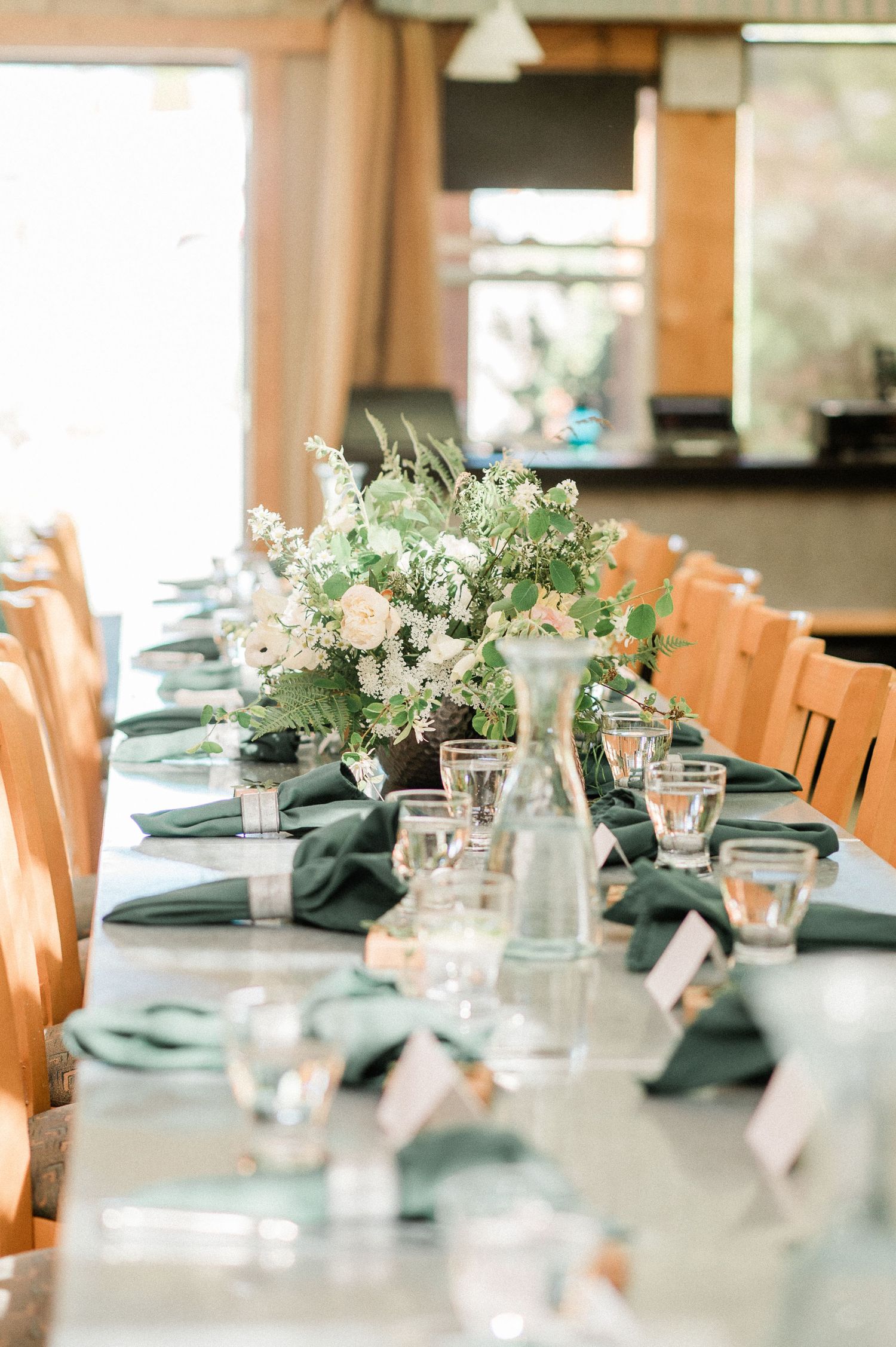 Elegant wedding reception table settings with white floral centerpieces, glass votives and sage green linens in natural light.