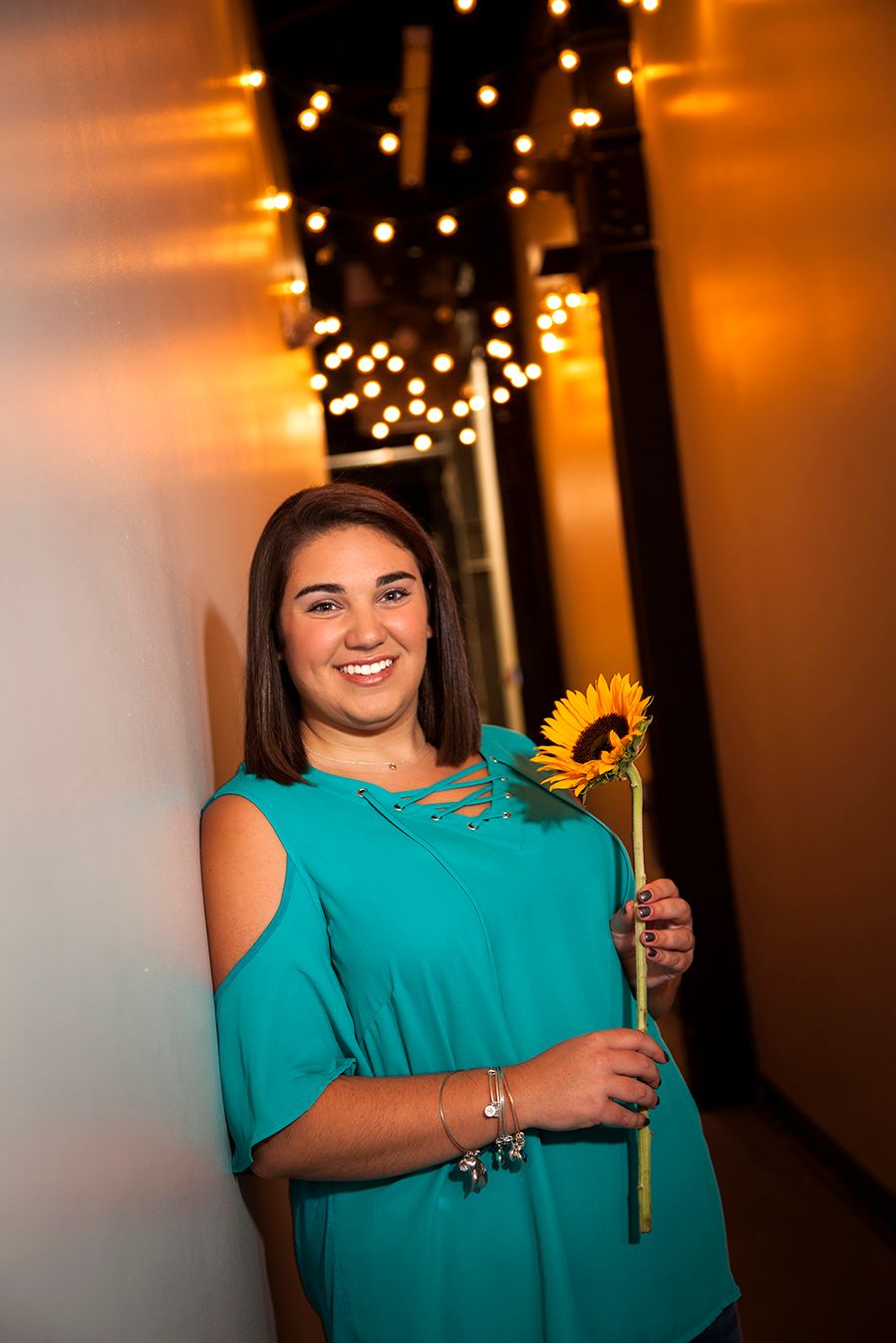 Someone in a turquoise dress poses with a sunflower in a dark hallway decorated with string lights. (Scottsville New York)