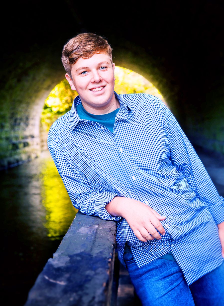 Someone in a blue checkered shirt and jeans poses along a stone wall near a tunnel with yellow lighting behind them. (Webster New York)