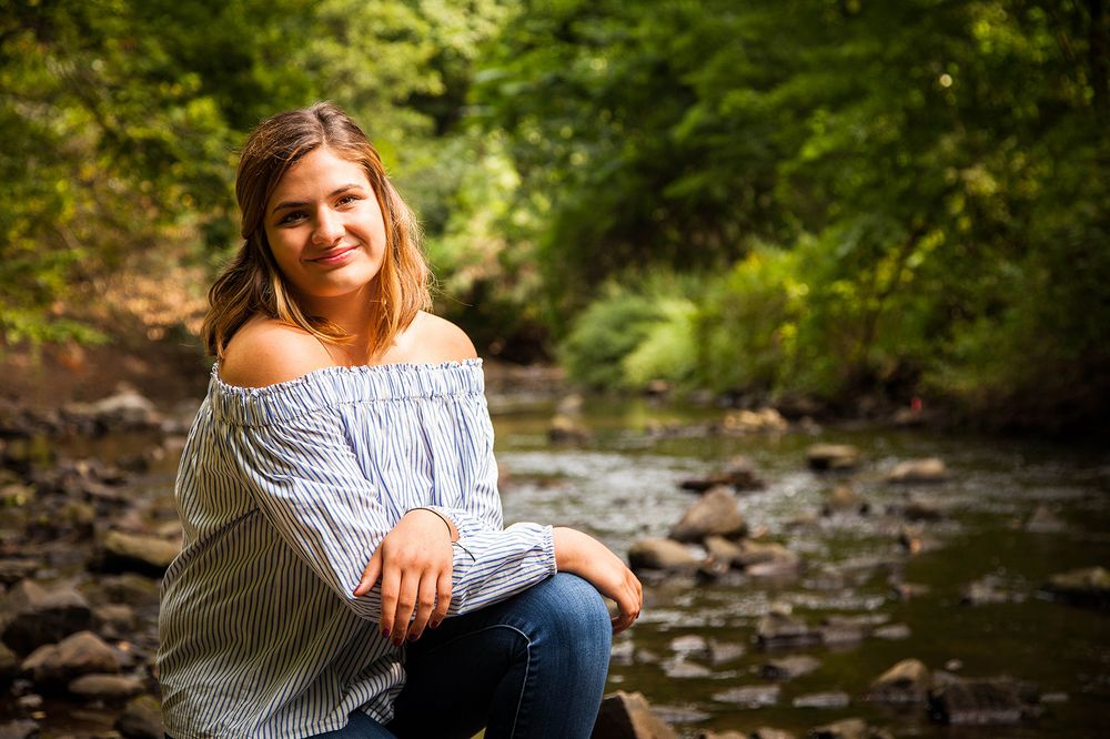 Someone in an off-shoulder white top poses near a rocky stream in a lush forest setting. (Holley New York)