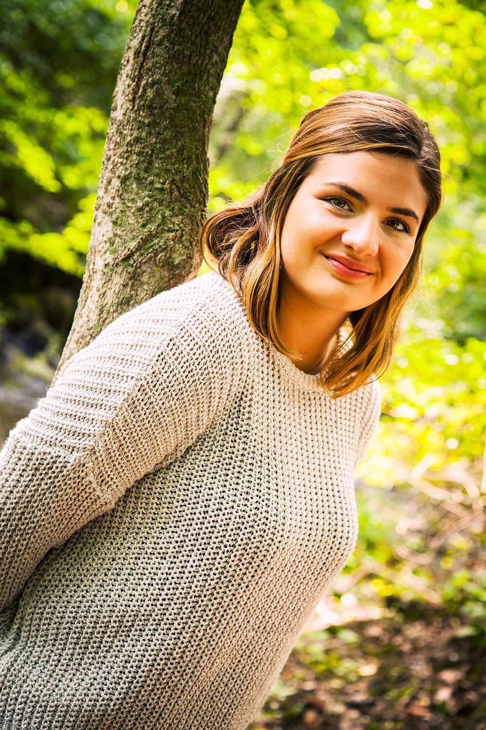 A person in a cream knit sweater stands beside a tree trunk in a sunlit forest with vibrant green foliage. (Kent New York)