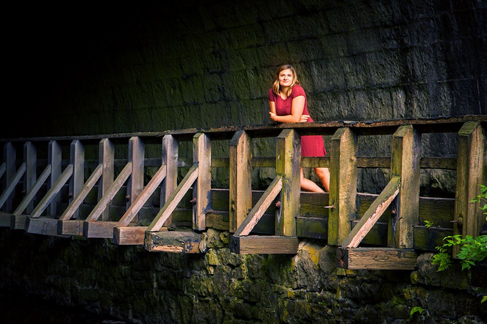 A girl in a red dress stands on a wooden bridge illuminated by dramatic lighting against a dark stone wall. (Corbett's Glenn Rochester NY)