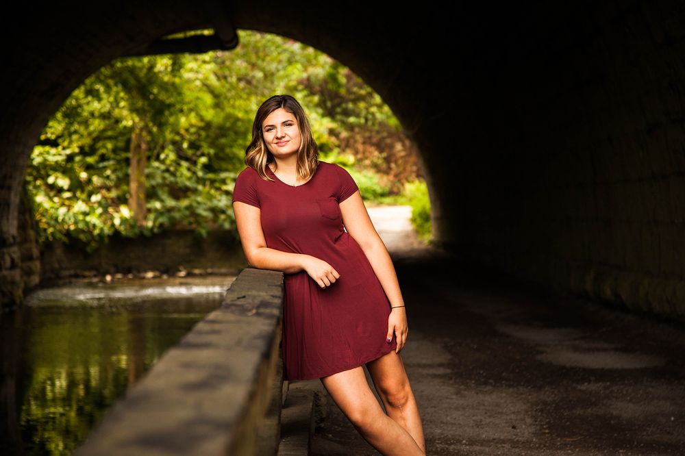 A girl in a red dress stands on a wooden bridge illuminated by dramatic lighting against a dark stone wall. (Corbett's Glenn Rochester NY)