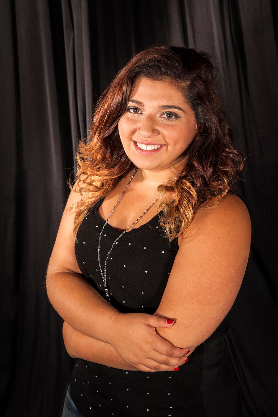 Portrait of someone with ombre curled hair wearing a black sequined dress against a dark curtain background, smiling warmly.