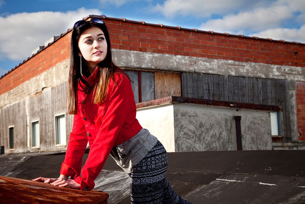 Someone in a bright red sweater leans against an industrial rooftop with dramatic lighting and urban architecture. (Hamburg New York)