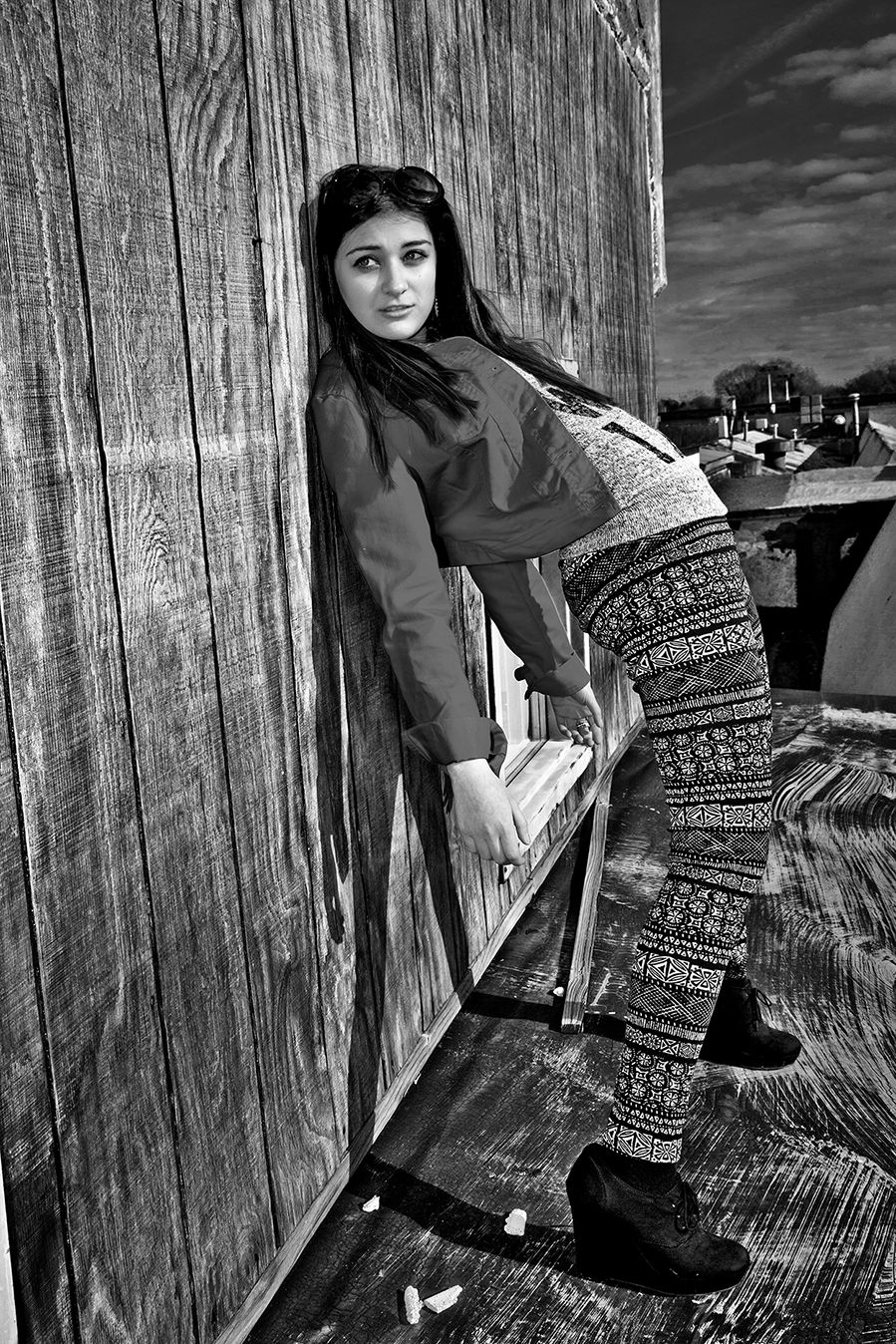 Black and white photo of a person wearing a knit cardigan and patterned pants leaning against a wooden wall outdoors. (Rush New York)