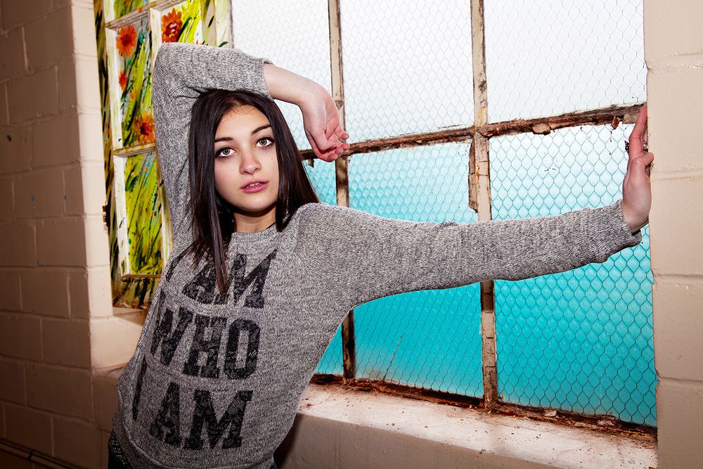 Figure in a gray hoodie sweatshirt poses by window with teal background in artistic photo shoot. (Mendon New York)