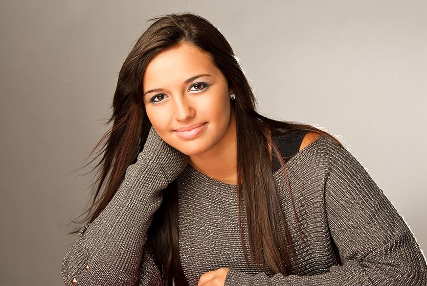 A smiling person in a cozy gray sweater leans on their hand while posing against a light neutral background.