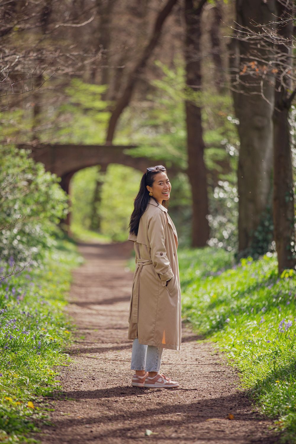 A woman in a beige coat walks along a wooded path lined with spring wildflowers and trees.