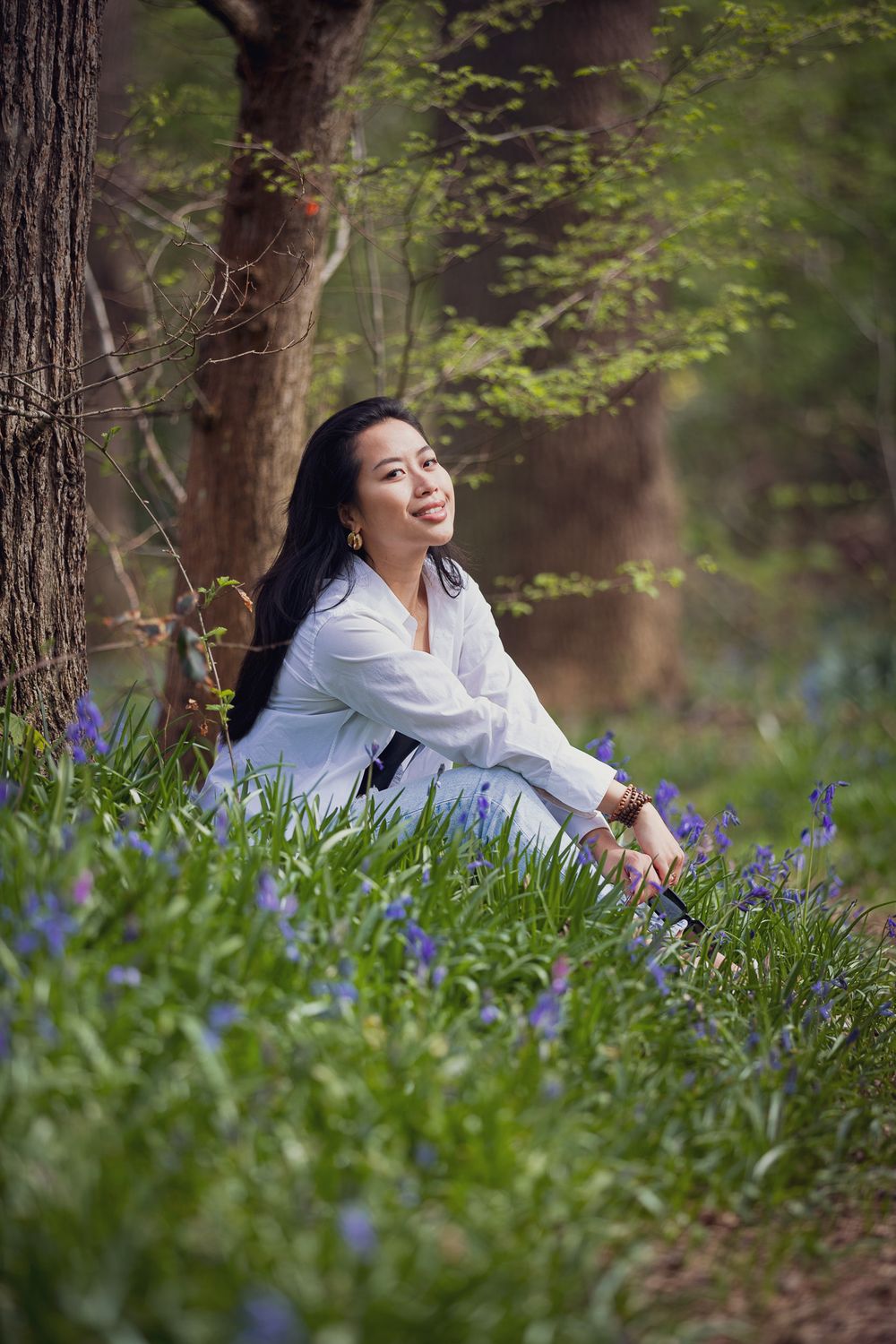 Woman in a white shirt sits peacefully among blooming bluebells in a tranquil forest setting.