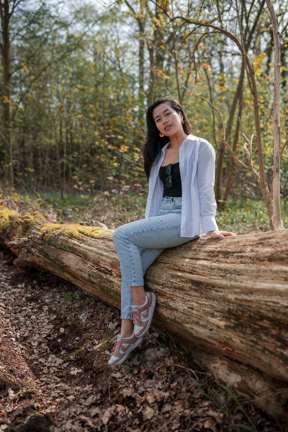 A person in white and denim sits casually on a fallen log in a wooded forest setting during autumn.