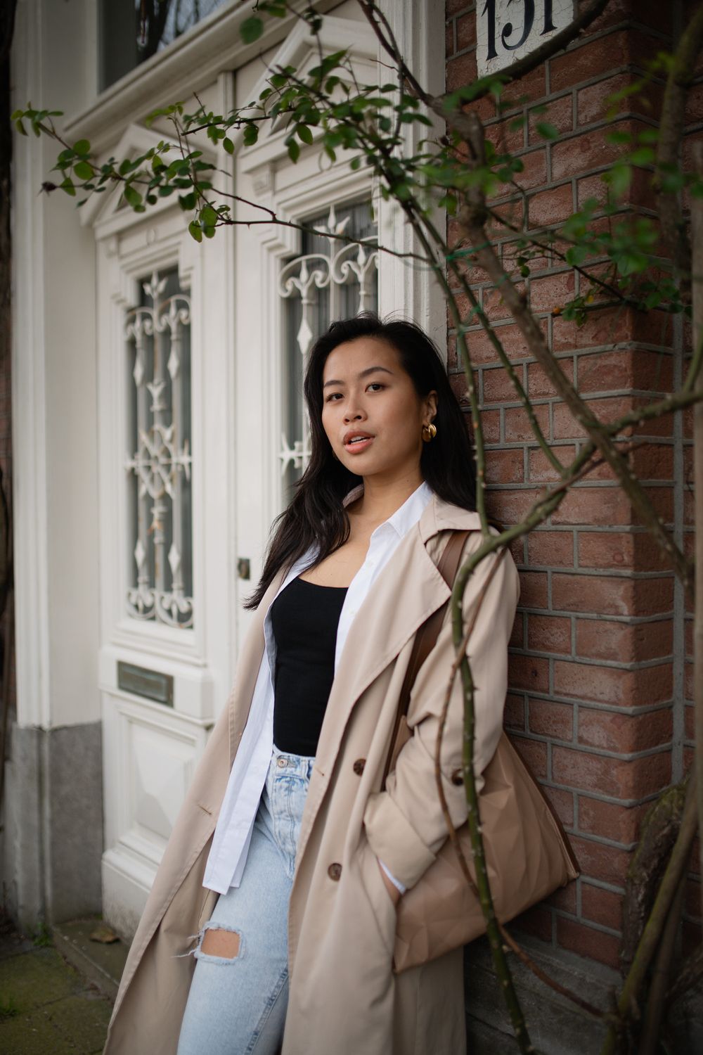A person in a beige trench coat and jeans poses against a brick wall with white windows in a fashion photoshoot.