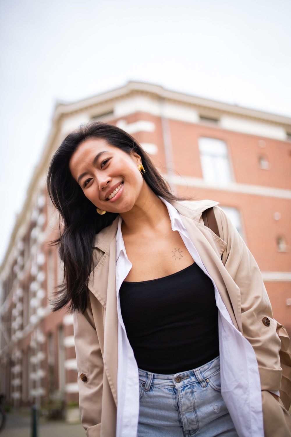 Woman in a beige trench coat and black top smiling playfully near brick buildings.