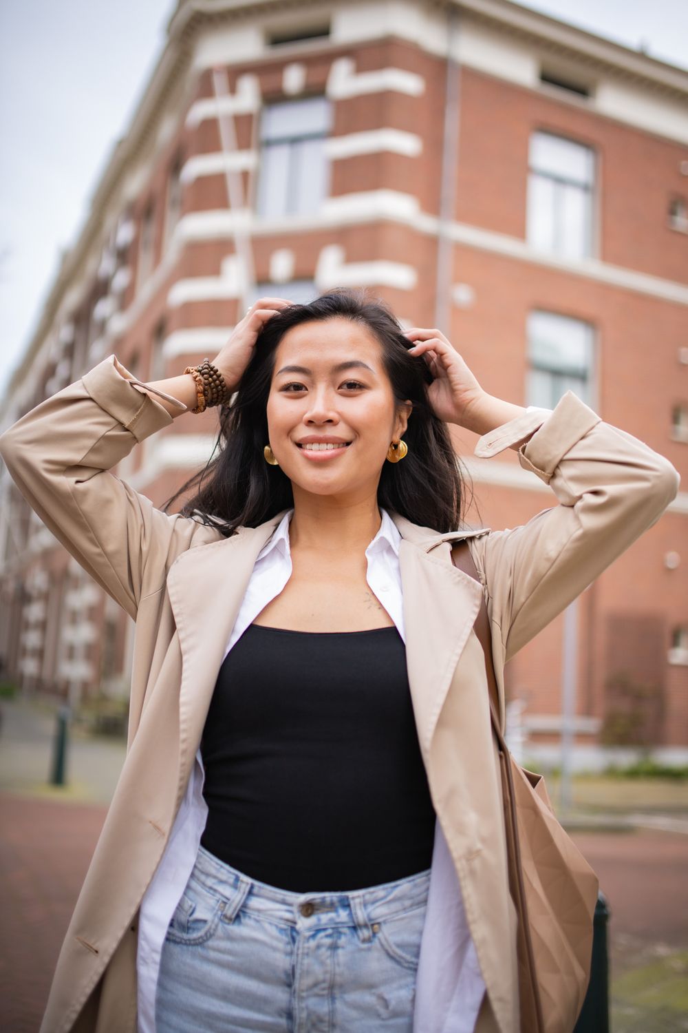 Woman in a beige trench coat and black top posing playfully outdoors near brick buildings.