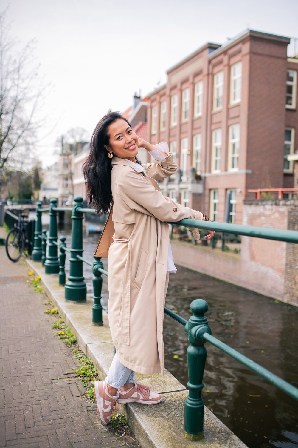 Woman in beige coat poses along canal railing in The Hague with brick buildings in background.