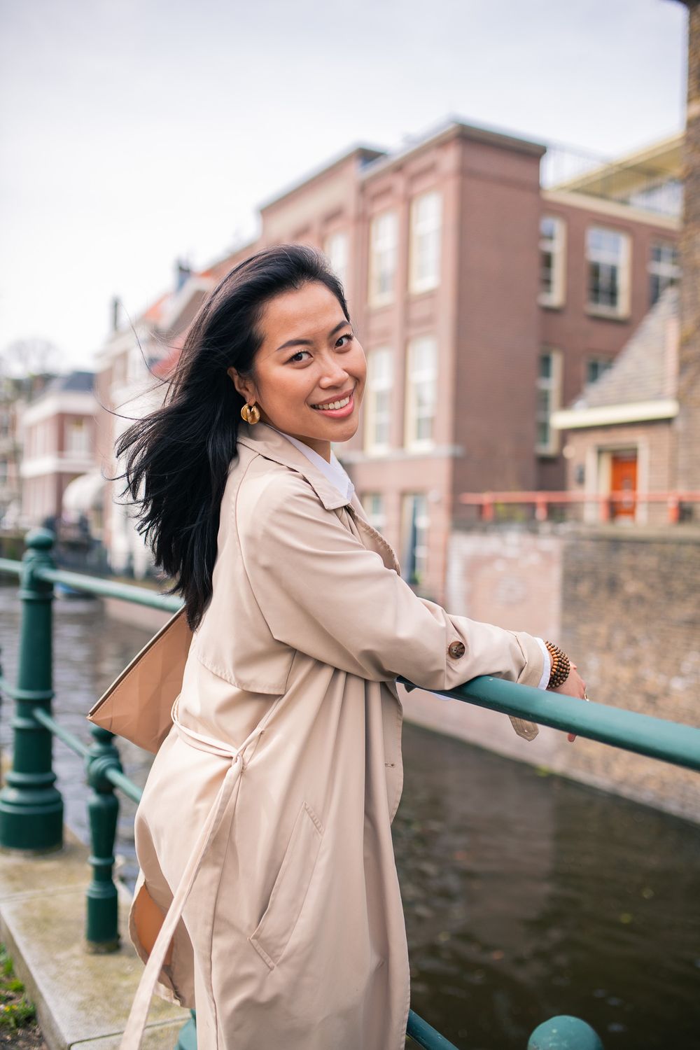 A person in a beige trench coat smiles while posing on a bridge in an urban setting.