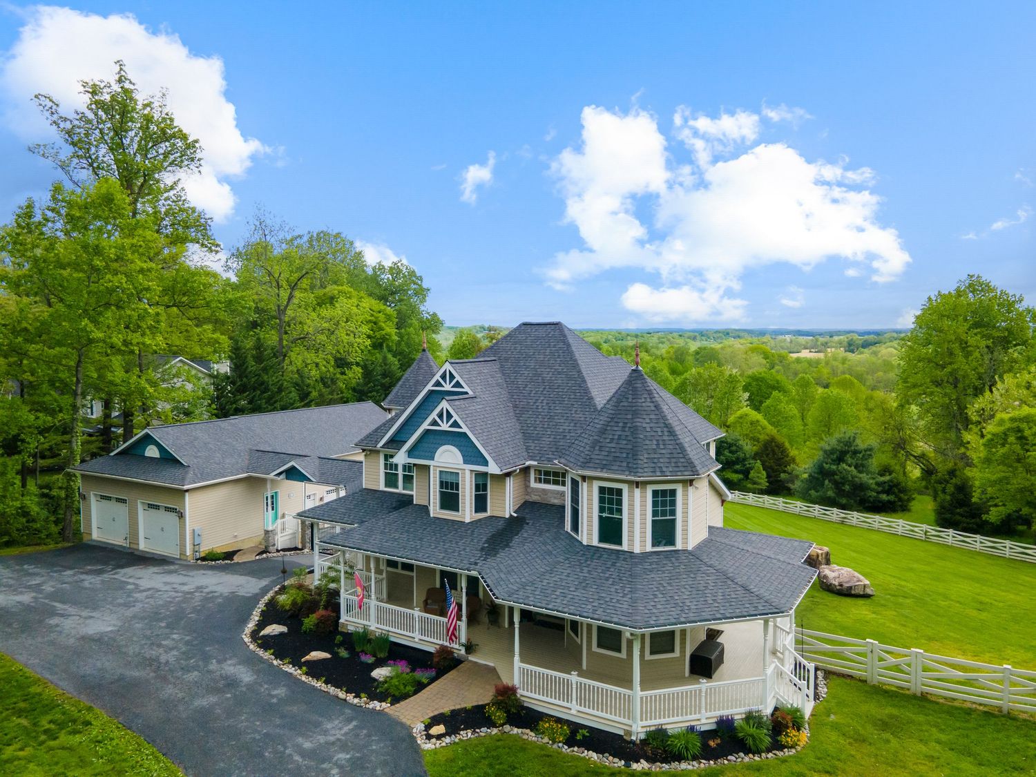 Aerial view of a luxury home with gray roof and wrap-around porch nestled in lush green surroundings.