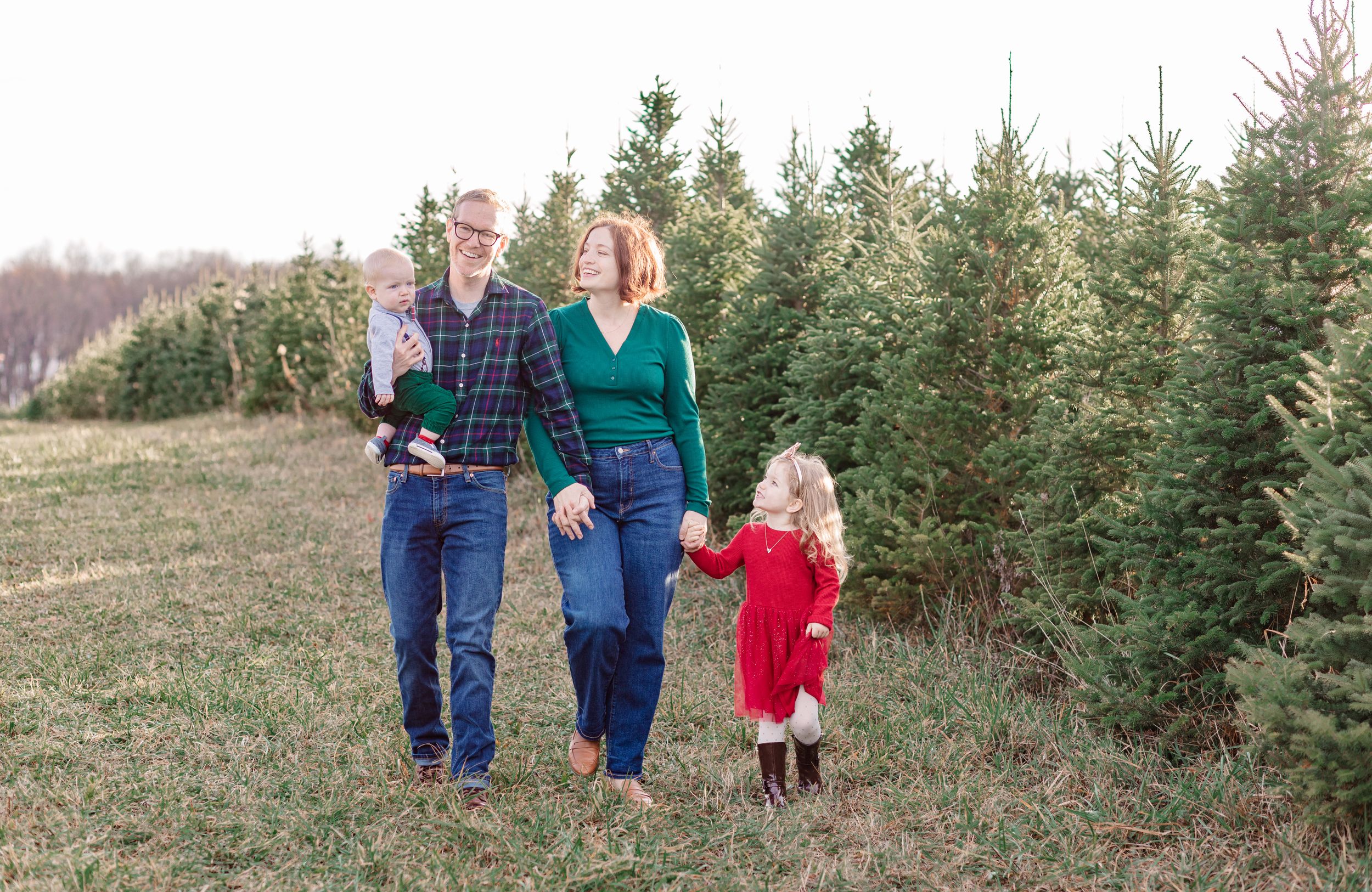 A family walks together through a Christmas tree farm with evergreen trees in the background. Family photography in Lansing, Michigan