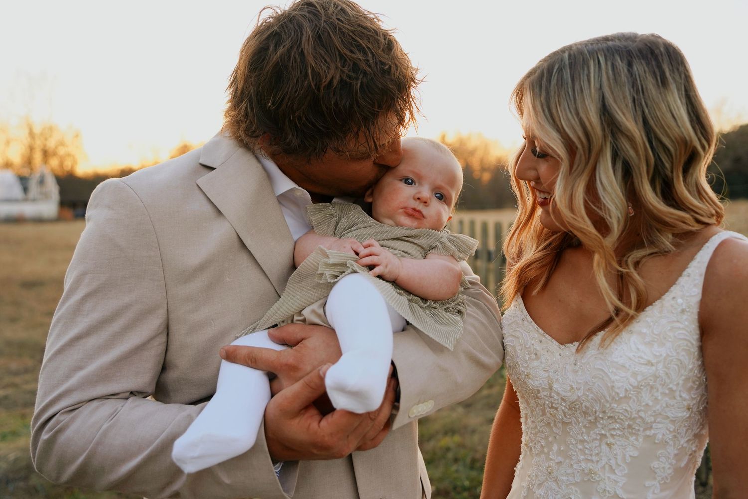 Bride and groom kissing newborn baby at golden hour during outdoor wedding celebration.