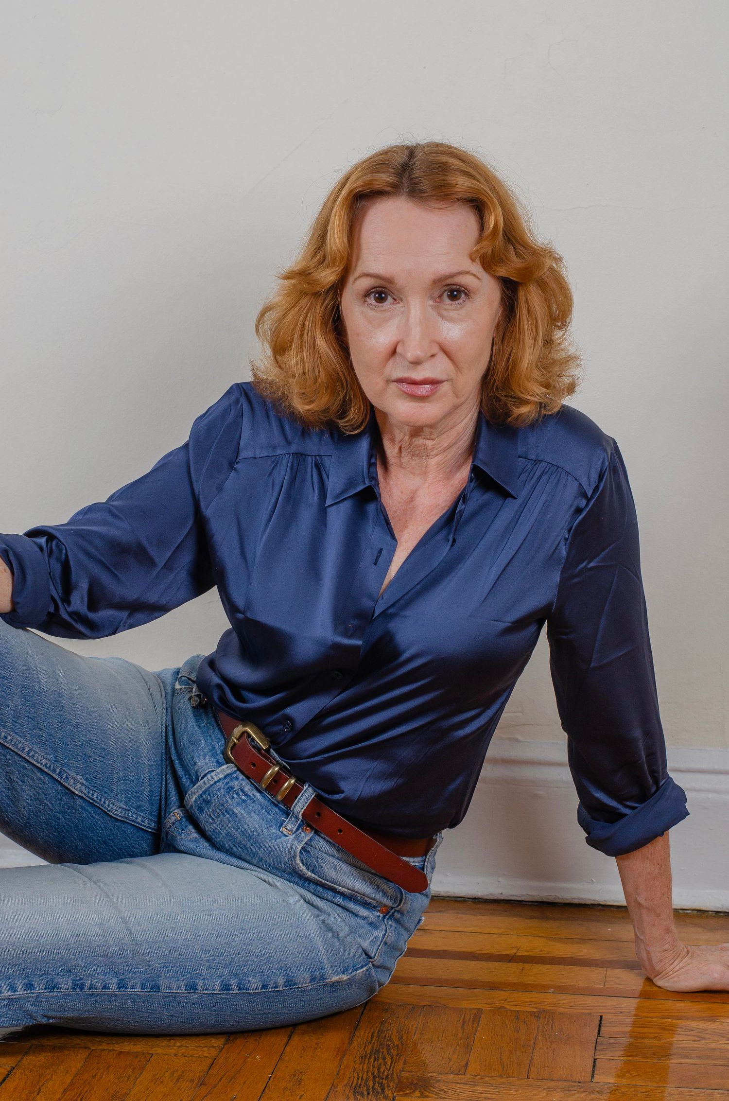 A woman in a navy silk blouse and jeans sits casually on wooden flooring.