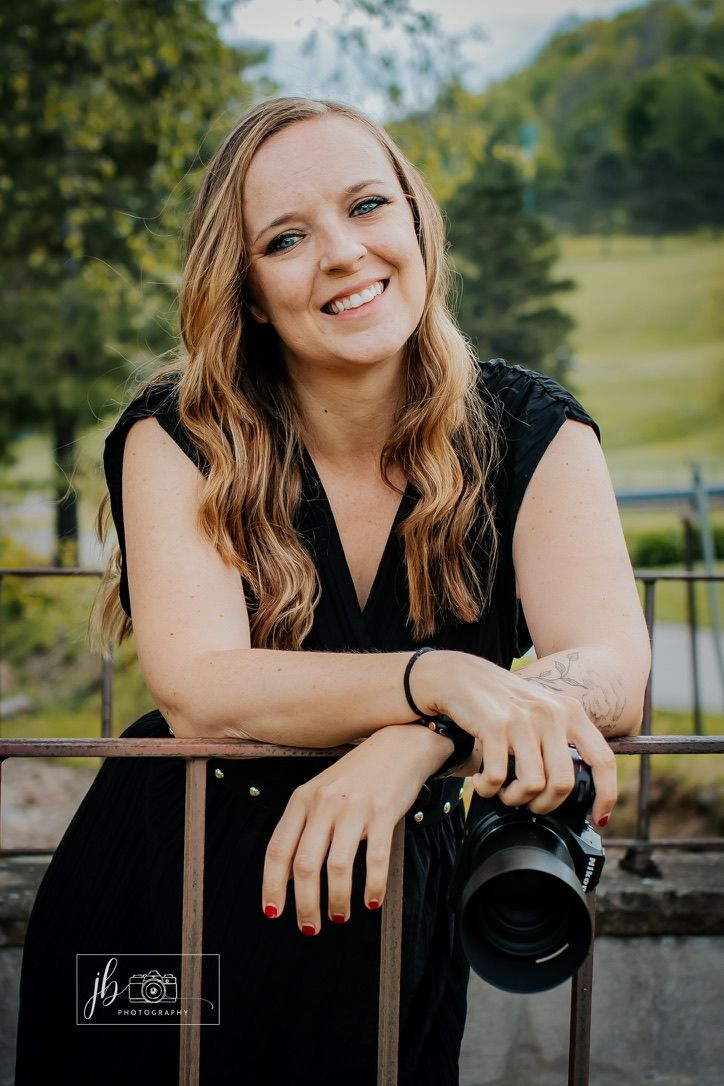 Professional photographer in black outfit poses with camera equipment in bright studio setting.
