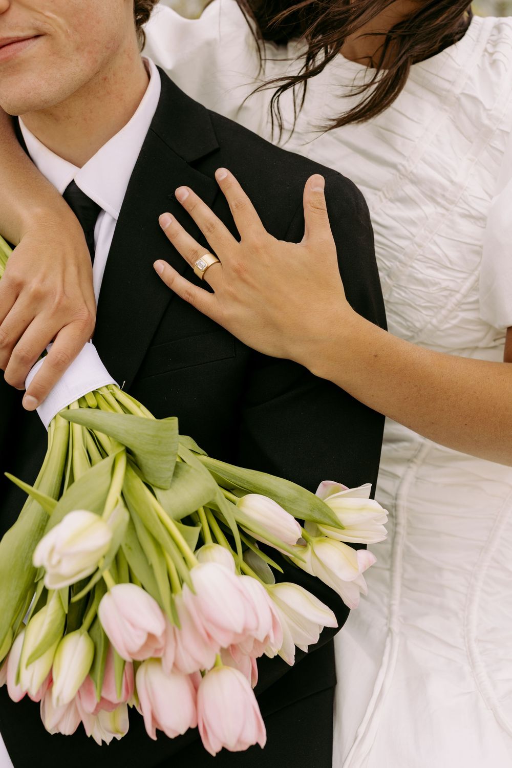 A close-up shot of a bride in white dress holding pink tulips while embracing a groom in black suit with tie.