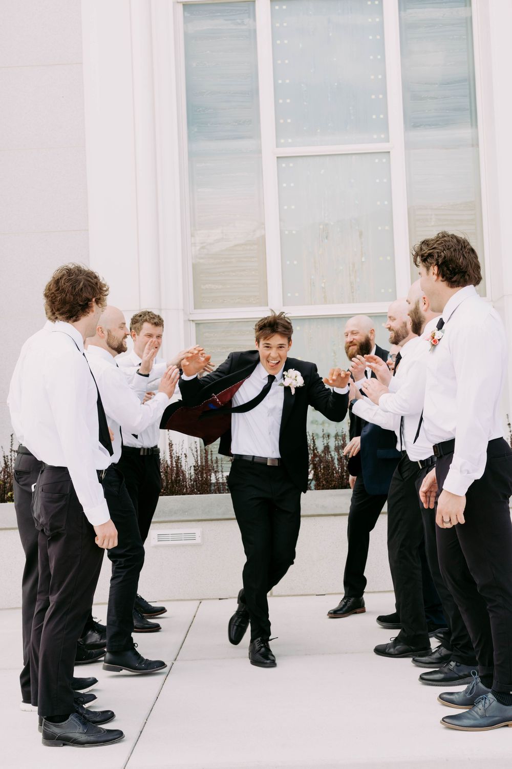 Groomsmen in black and white formal attire form a line while one jumps and dances in celebration outside a building.