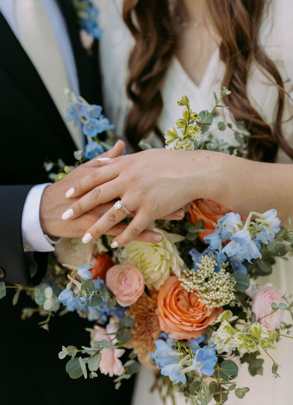 Close-up of hands with wedding ring holding beautiful bouquet of roses and blue delphiniums with eucalyptus accents.
