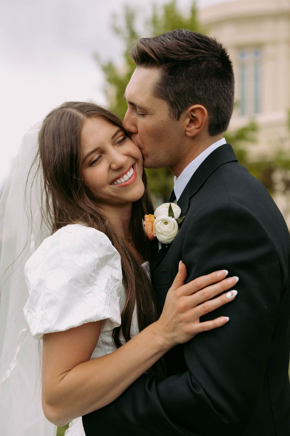 A joyful bride in a white dress with puff sleeves shares an intimate moment with her groom in a black suit outdoors.