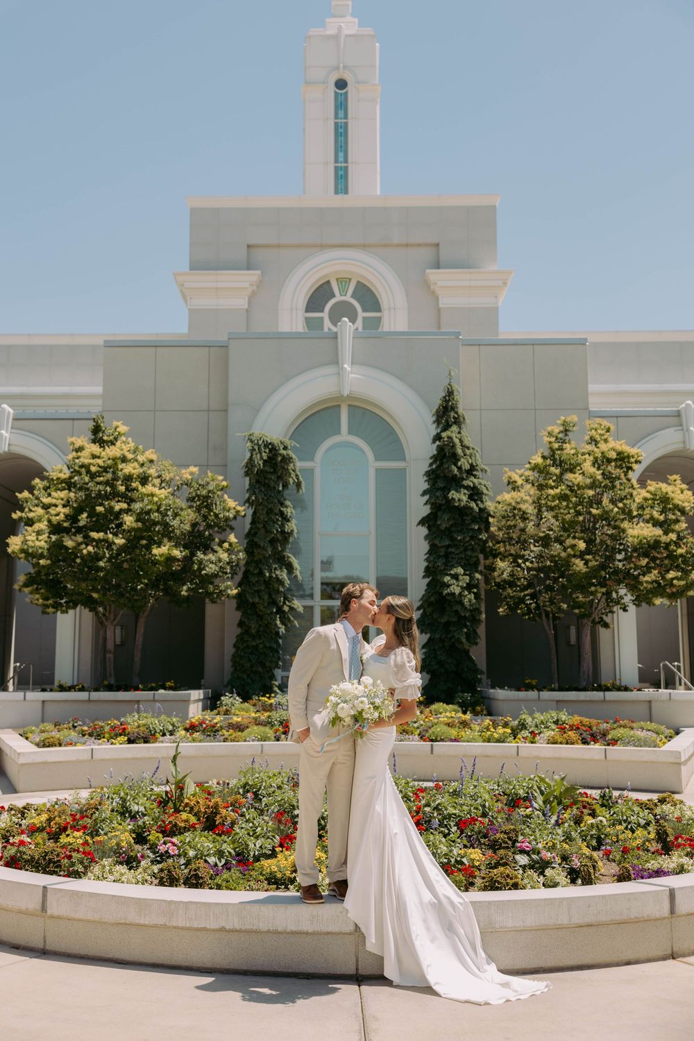 A couple stands together in formal attire outside a Mormon temple with white architecture and flowering gardens.