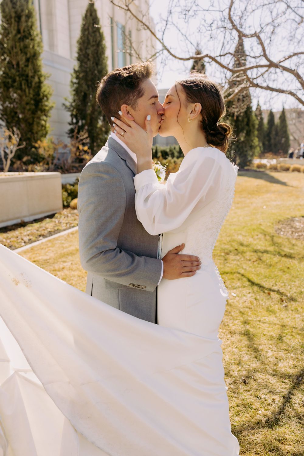A couple shares a tender kiss on their wedding day outdoors near evergreen trees in spring.