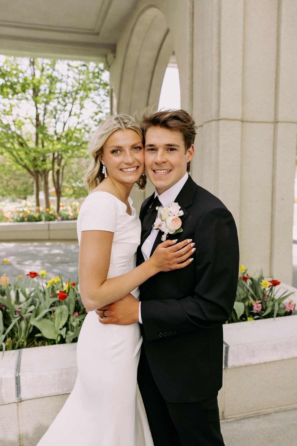 A couple poses together on their wedding day wearing traditional white dress and black tuxedo near blooming tulips.