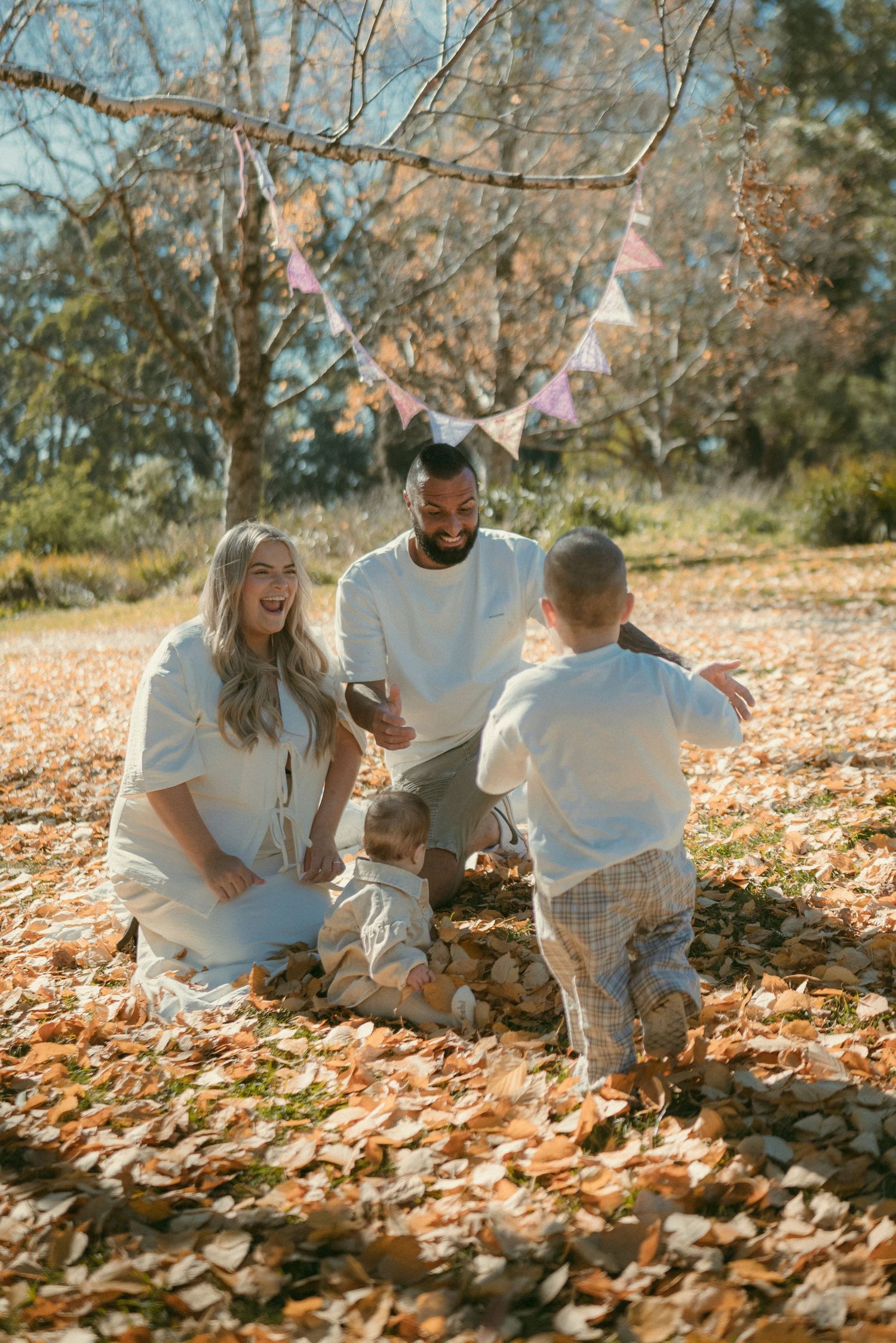 Family laughing and playing in autumn leaves outdoors, with bunting decorations hanging from trees on a sunny day.