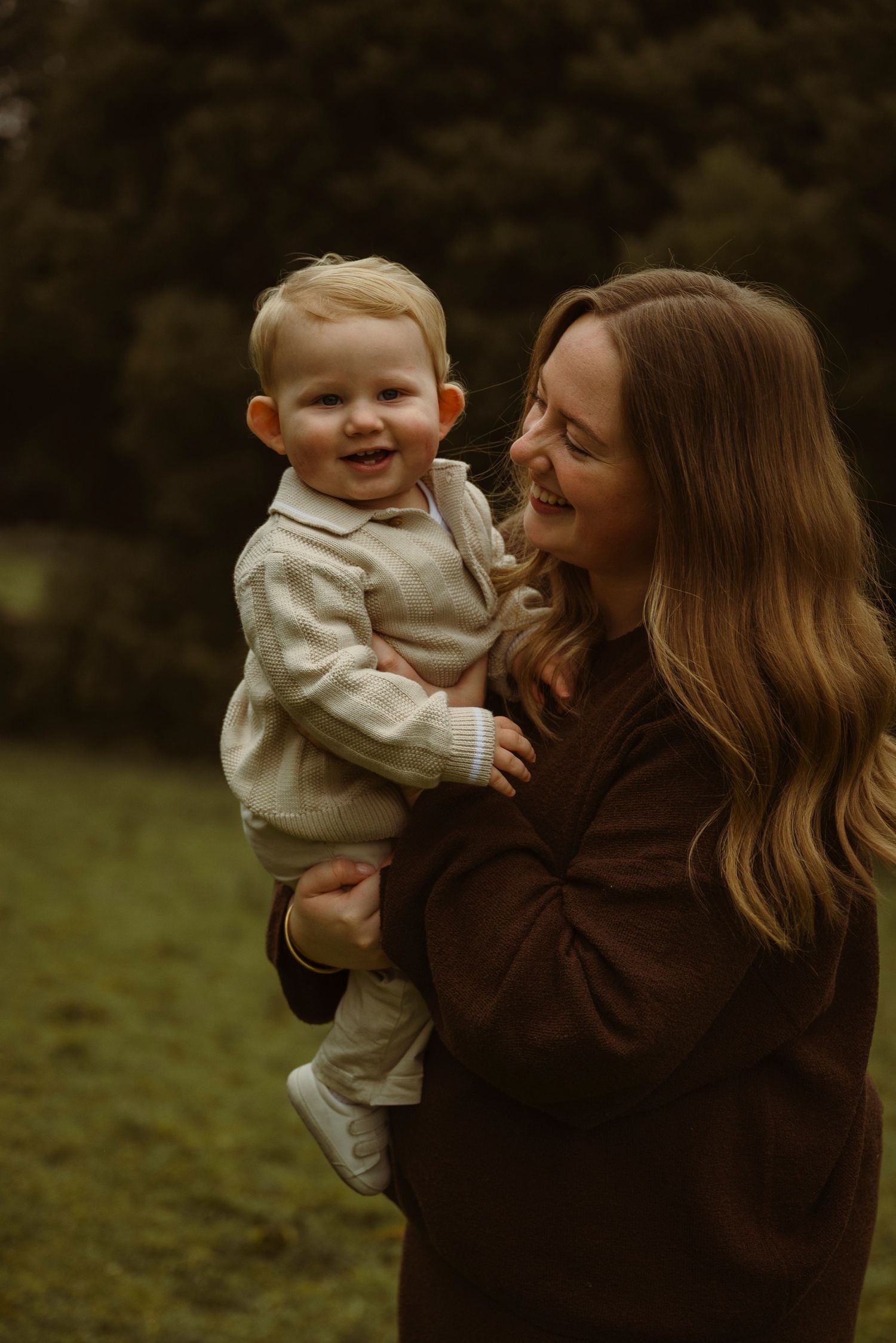 A smiling mother in a brown jacket holds her happy toddler boy outdoors in a lush green park setting.