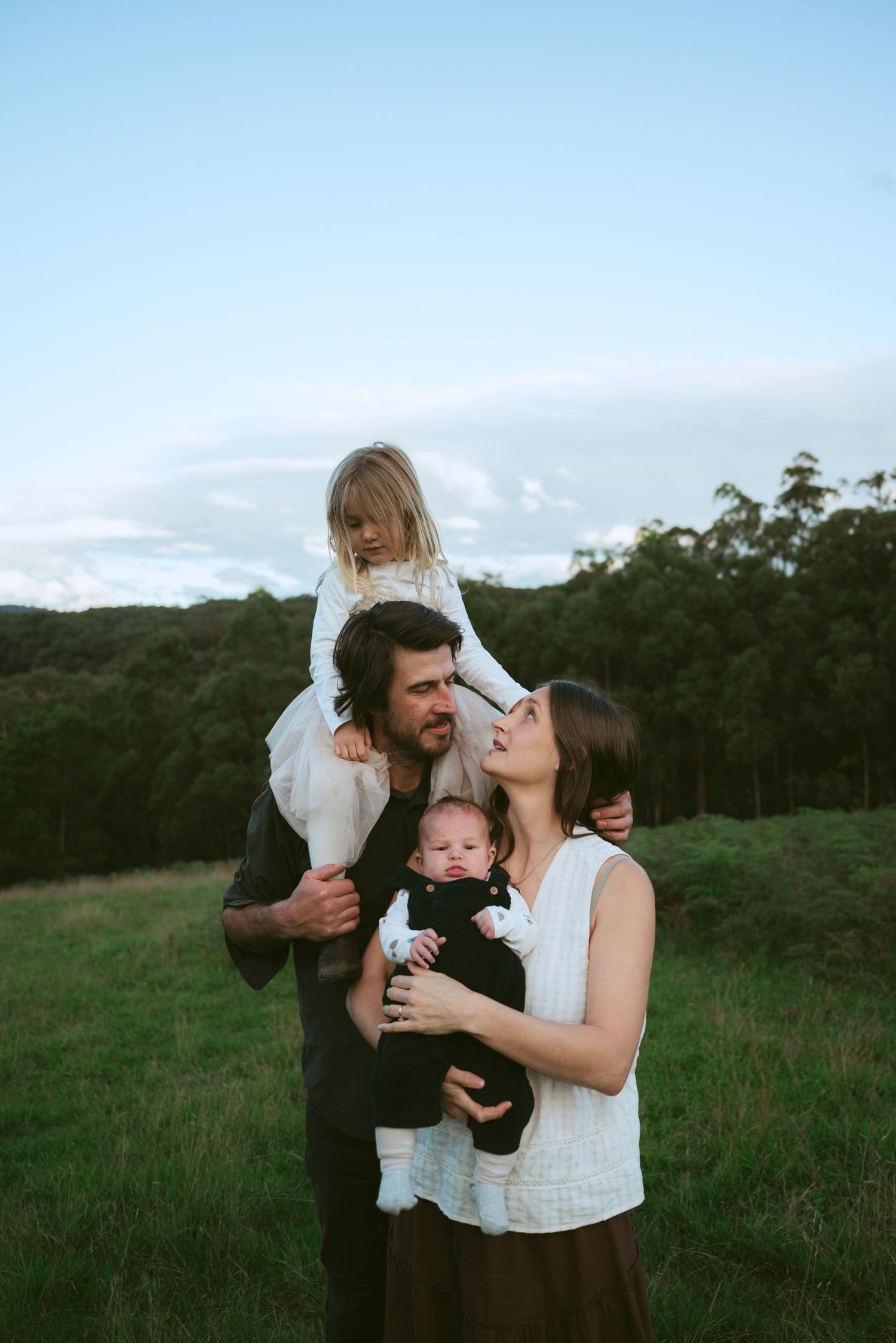 Family of four poses outdoors on green hillside, father holding toddler on shoulders and mother cradling newborn baby.
