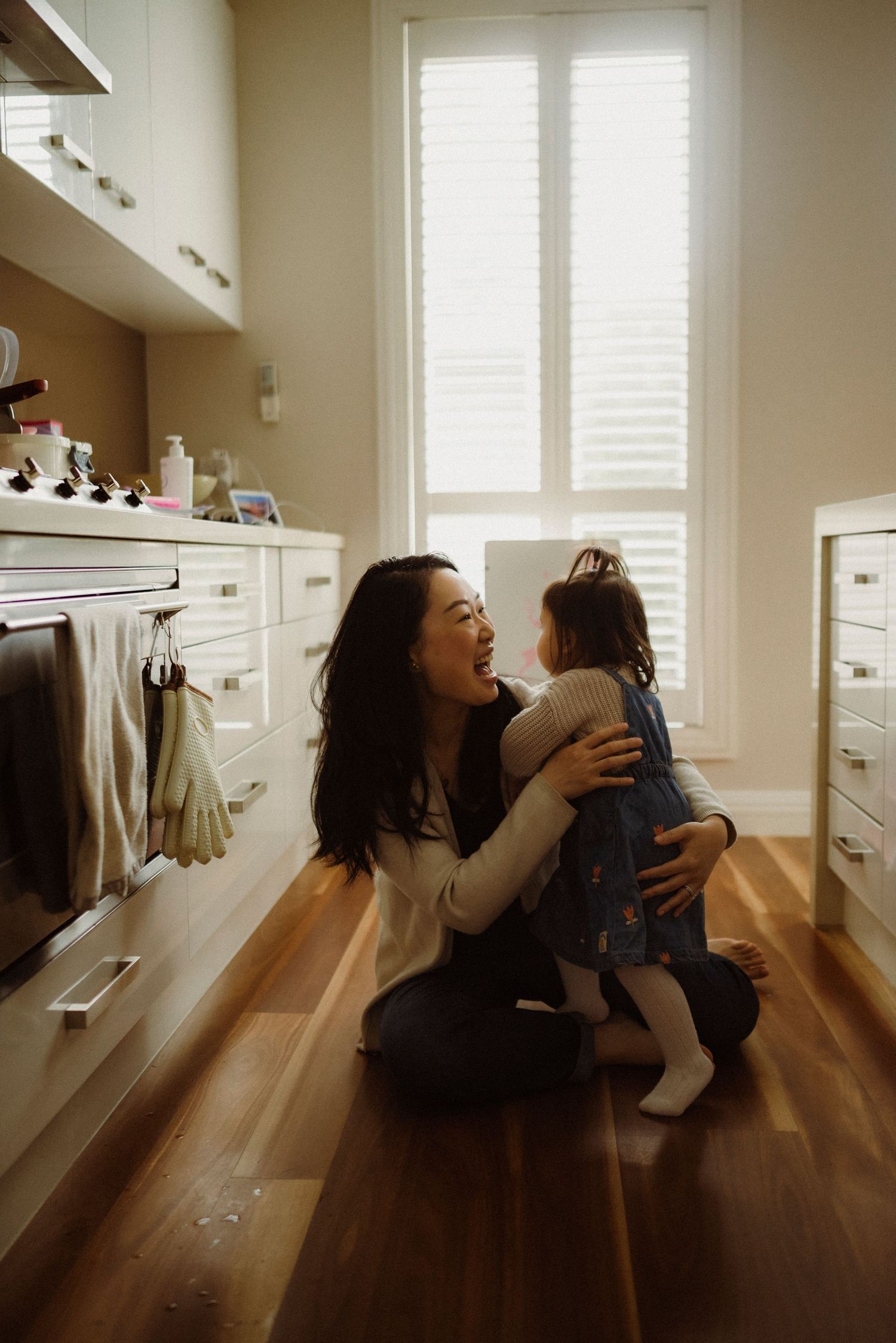 A mother kneels on hardwood floor hugging her toddler daughter in a bright kitchen, sharing a joyful moment.