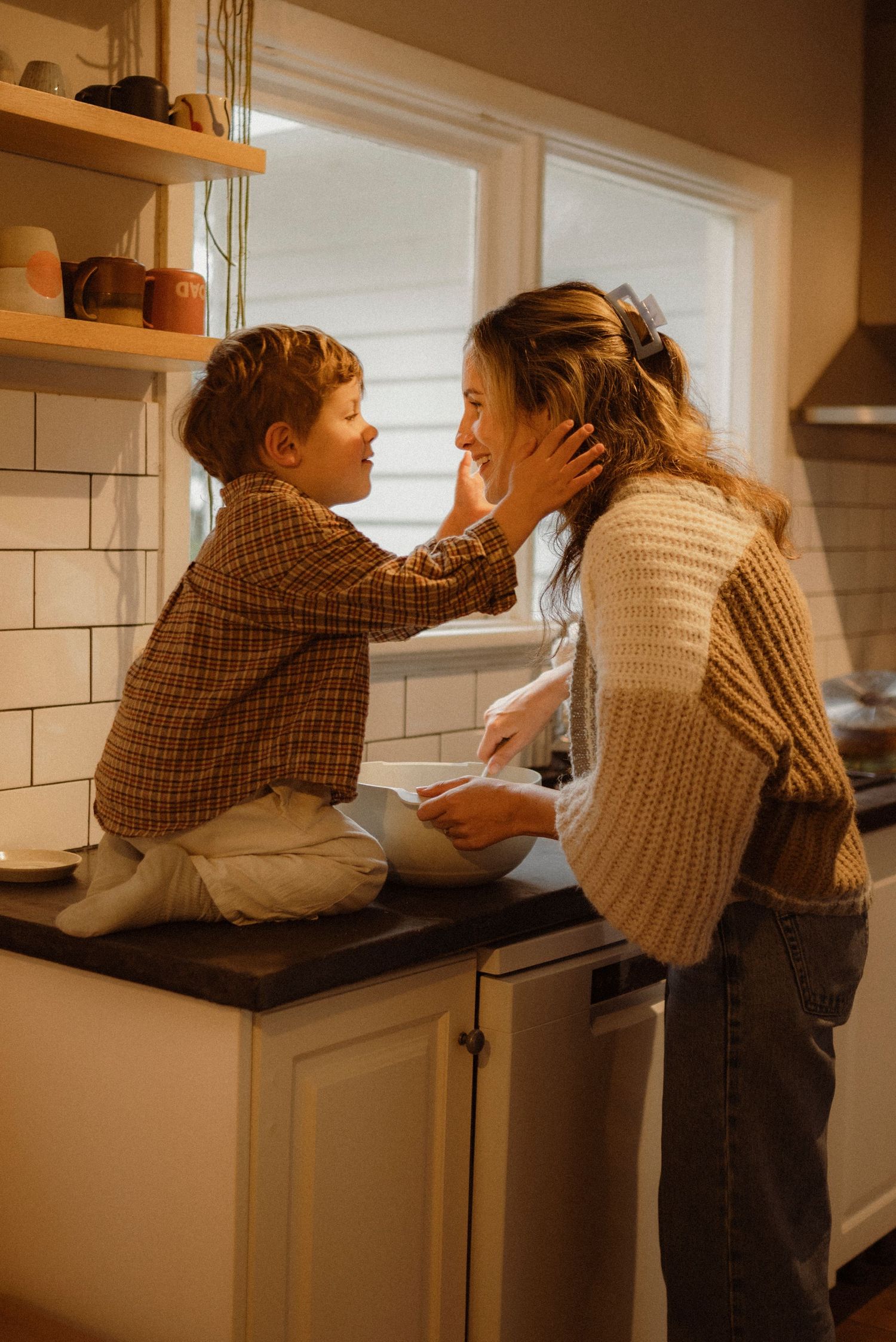 A young child sits on a kitchen counter, tenderly touching a woman's face while she holds a bowl in a warmly lit home.