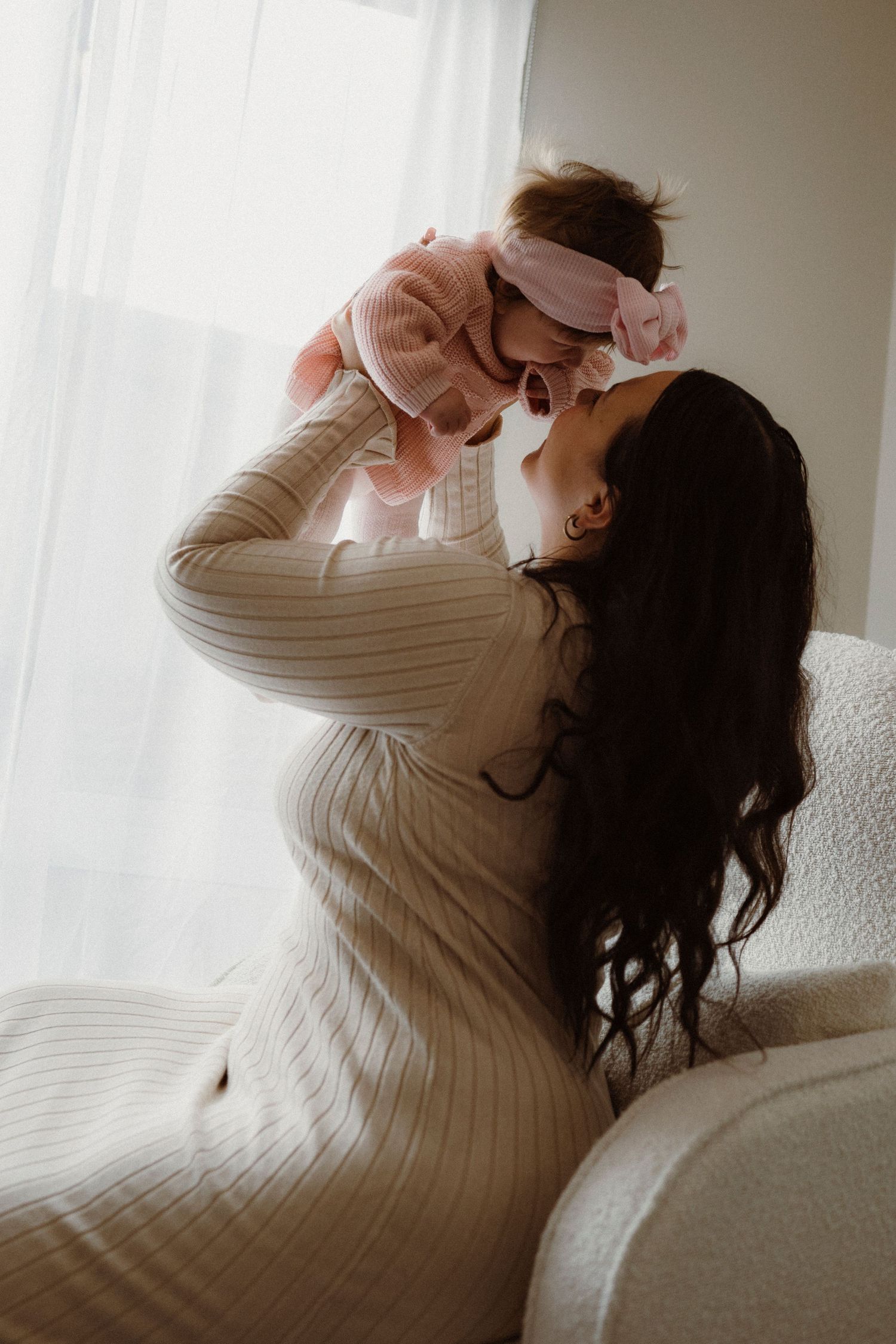 A mother in a cream ribbed dress lifts her baby in pink outfit near a bright window.
