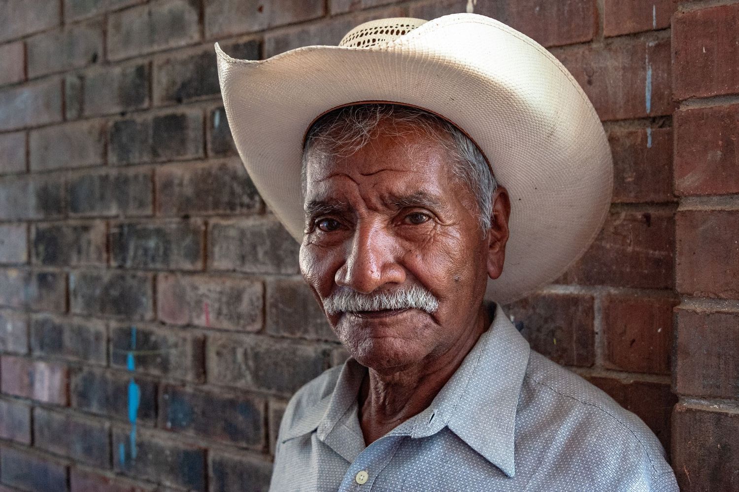 A person in a cowboy hat smiles while sitting against a rustic brick wall background.