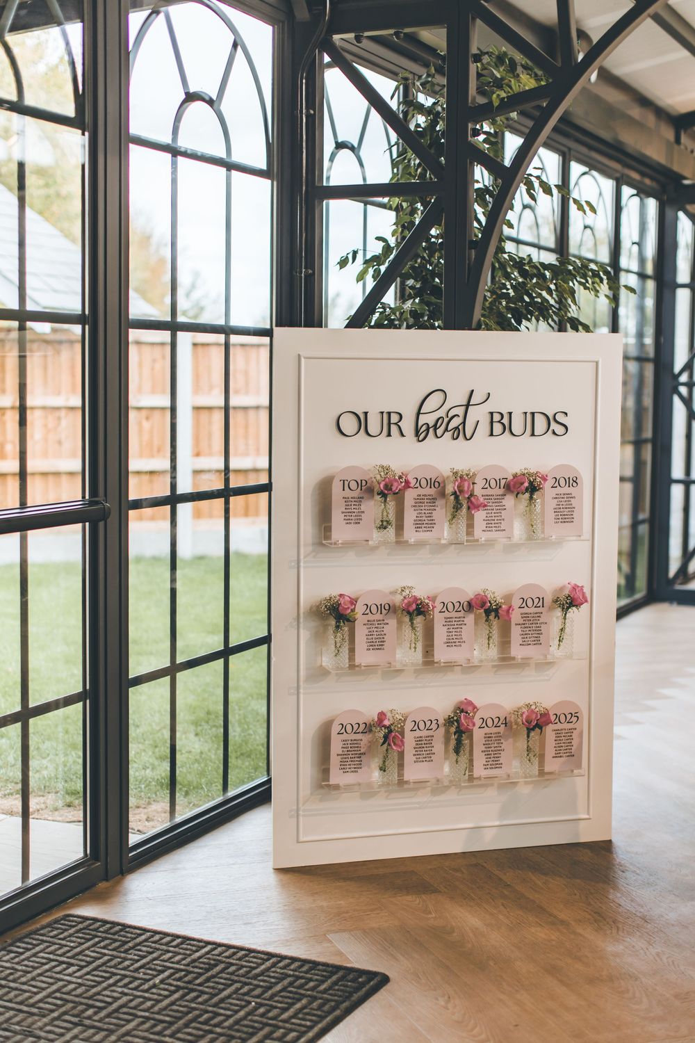 Wedding seating chart display titled 'Our Best Buds' with floral decorations in a sunlit venue.