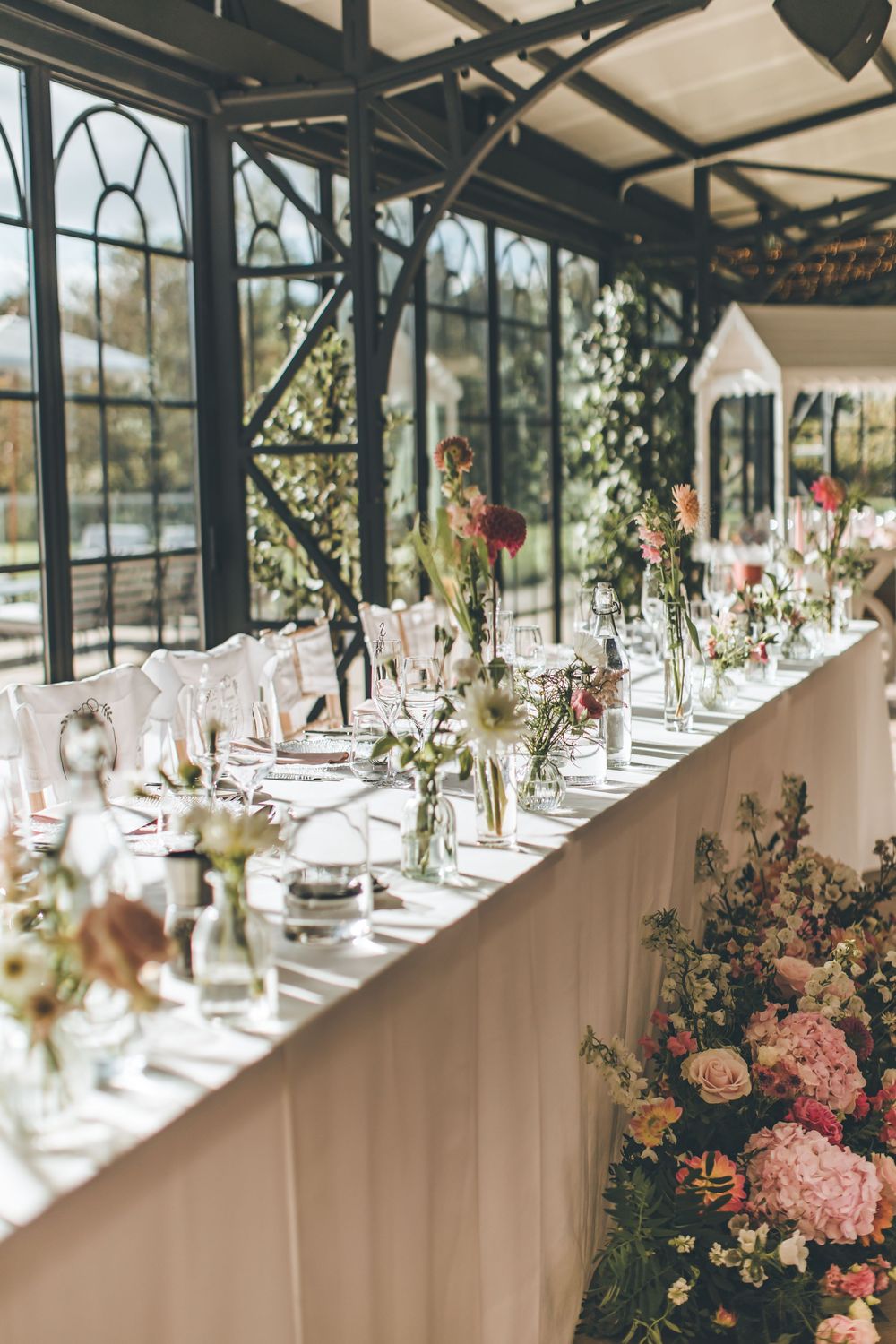 Elegant wedding reception table setting with delicate floral centerpieces in a sunlit greenhouse venue.