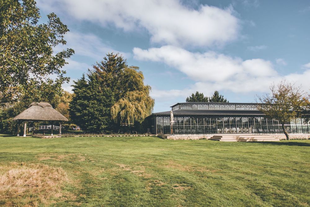 Modern event venue building with glass walls surrounded by green lawn and trees under blue sky.
