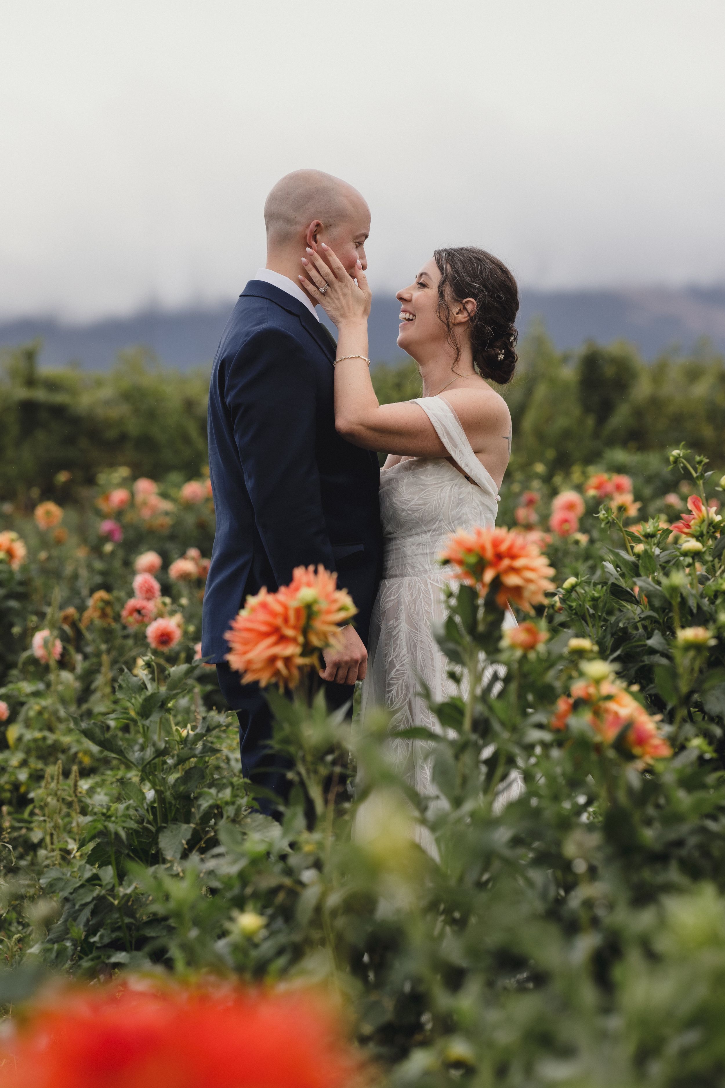 A romantic wedding couple shares an intimate moment surrounded by blooming orange dahlias in a garden setting.
