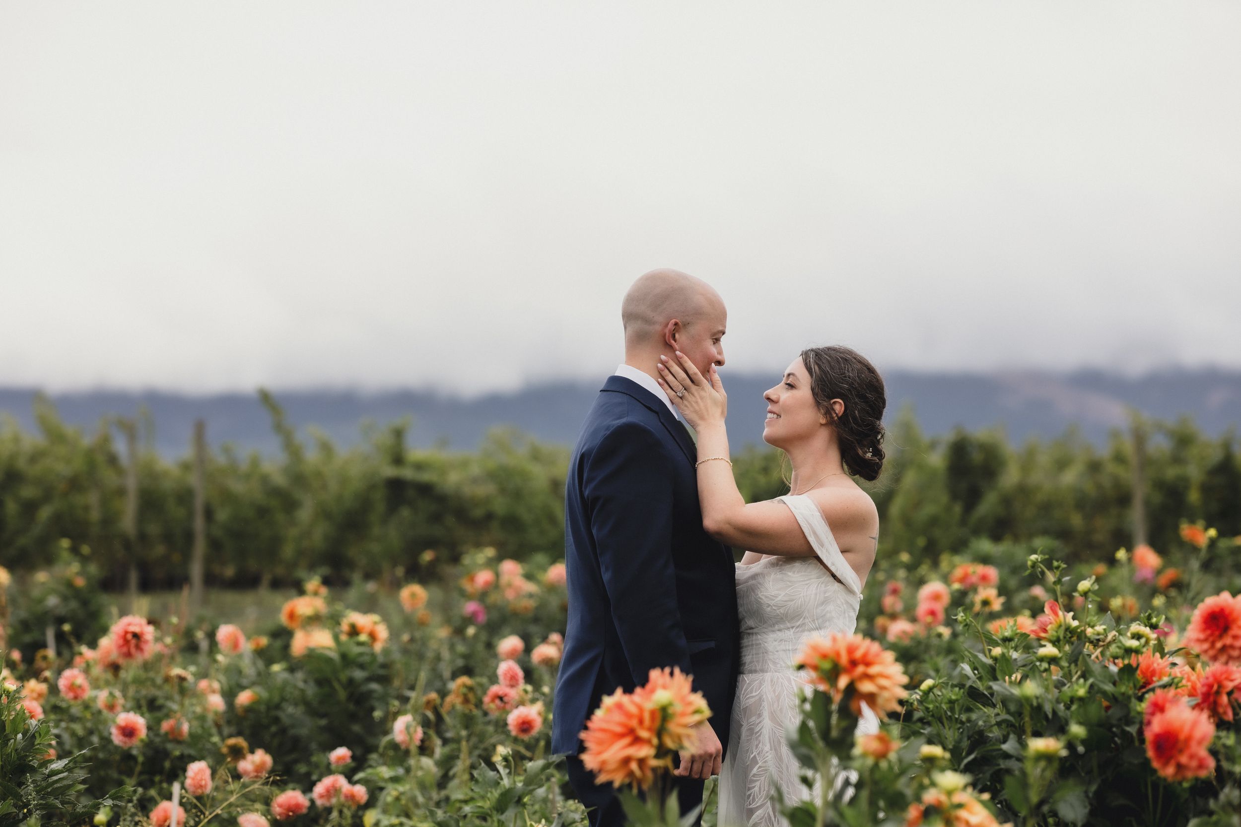 A couple shares a romantic moment in a blooming dahlia flower field with mountains visible in the distance.