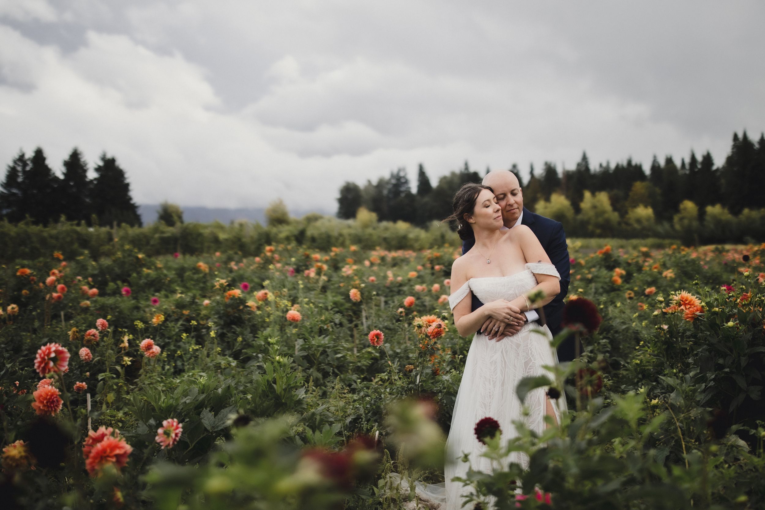 A couple poses together in a vibrant flower field filled with orange and pink blooms on an overcast day.