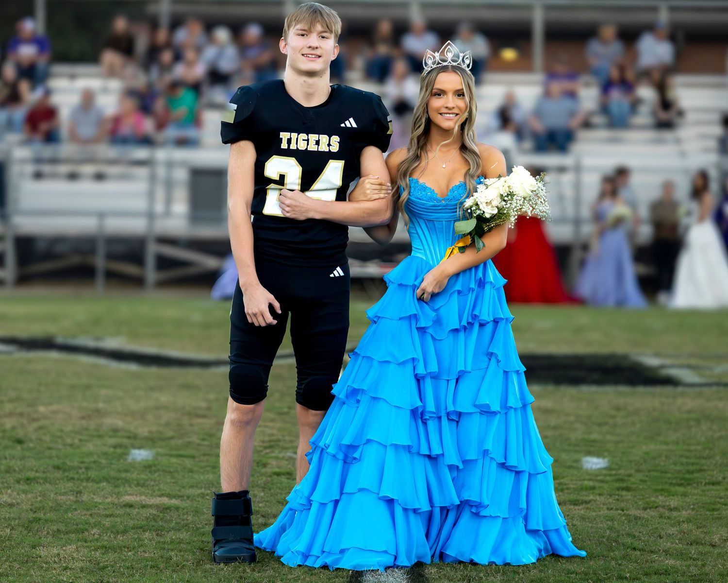 Two students pose together on a football field, one in a black football uniform and one in a bright blue formal gown.