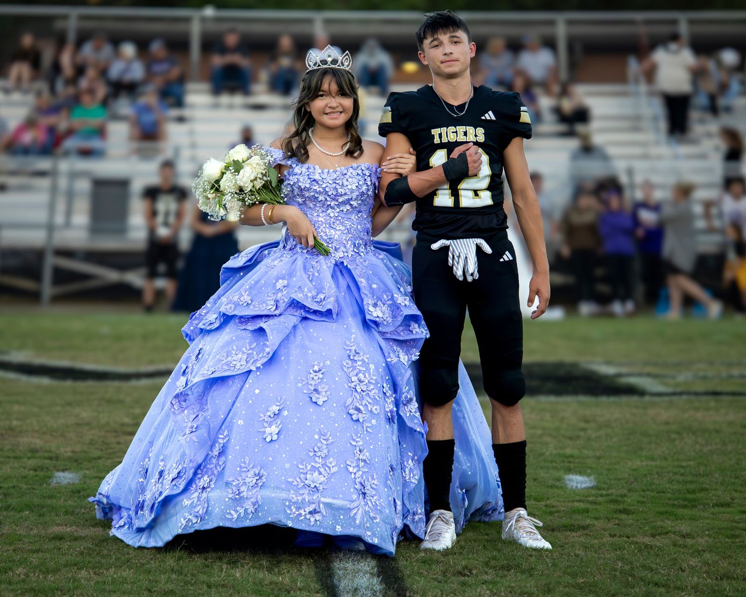 Children in formal attire, including a light blue gown and football uniform, walk together on a grassy field.