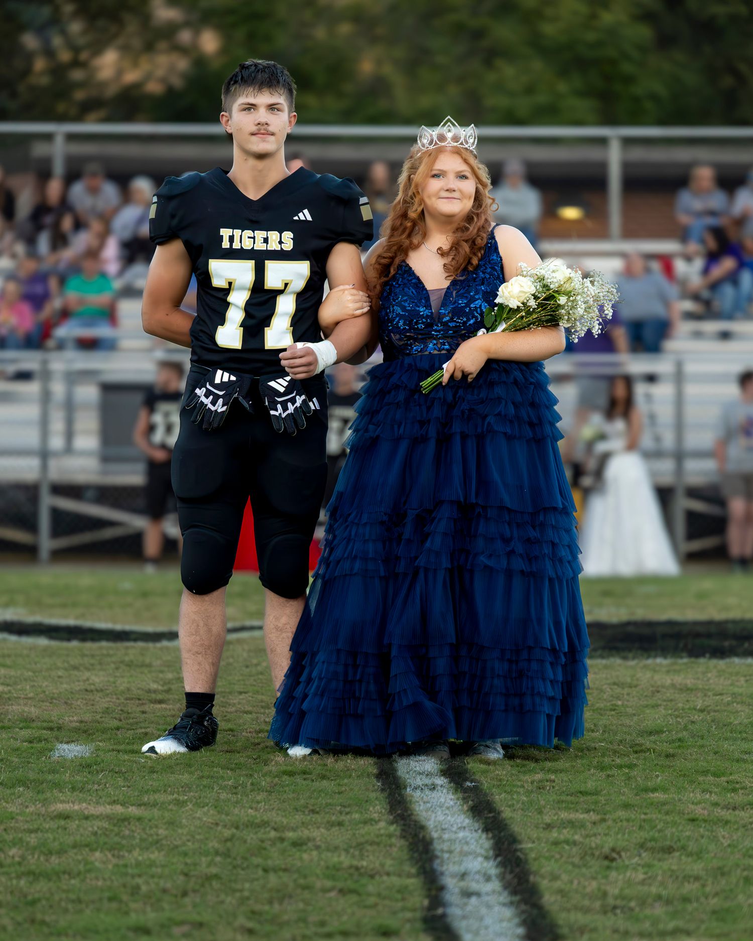 A football player in a Tigers jersey stands with a homecoming attendant in a navy blue gown on the field.